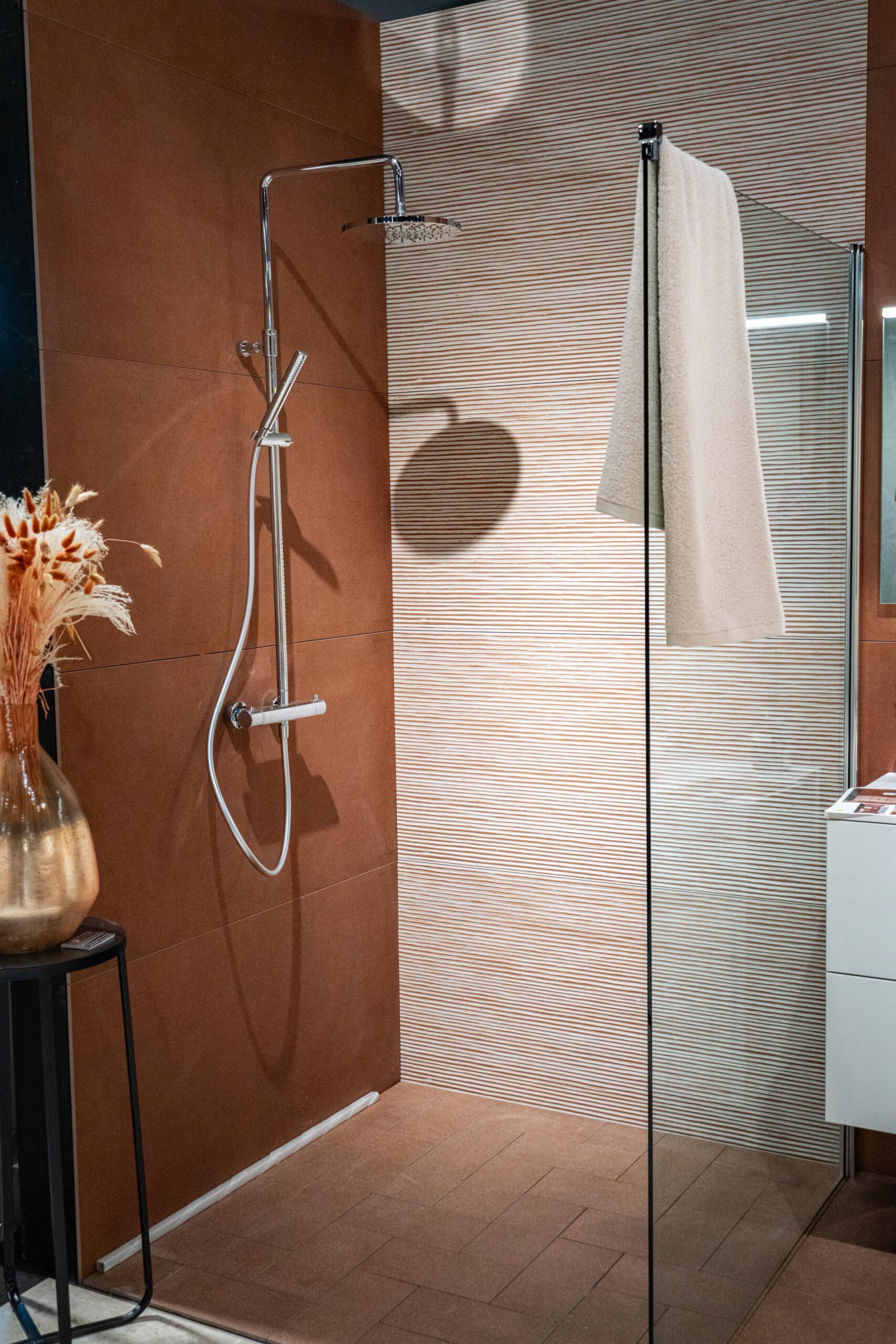Douche chromée devant un mur terracotta et un panneau clair ondulé, avec paroi vitrée.